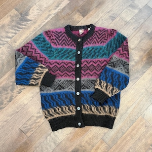 May include: A patterned cardigan in shades of pink, teal, blue, grey, and tan. The button-up sweater has a black collar and cuffs. The geometric design features horizontal stripes. It has six white buttons.