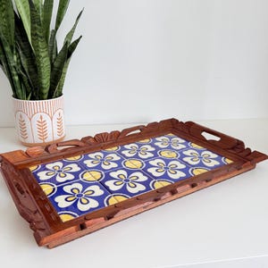 May include: A rectangular wooden serving tray with a decorative tile insert. The tiles feature a blue background with white and yellow floral patterns. The tray has carved handles and edges, and is placed on a white surface.