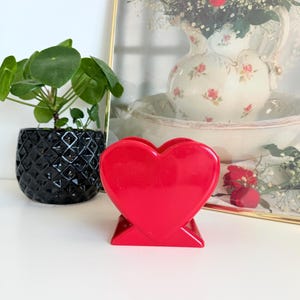 May include: A vibrant red heart-shaped ornament is displayed on a white surface. A small potted plant with round green leaves is to the left in a black geometric pot. A framed picture of roses is in the background. The object is likely a decorative item or holder.
