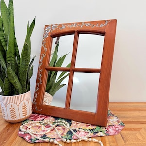 May include: A wooden mirror with a window pane design. The frame is a warm brown color, with floral and leaf accents. The mirror is resting on a floral patterned cloth, with a pearl necklace draped across it. A potted plant is in the background.