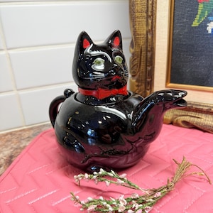 May include: A black ceramic teapot shaped like a cat with a red bow tie. The cat teapot has green eyes and red accents on its ears. The teapot is sitting on a pink quilted mat with a small bundle of flowers.