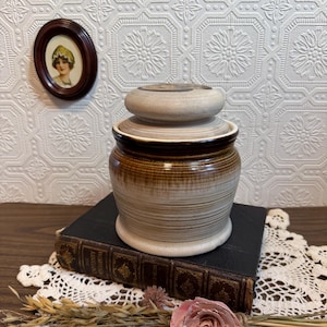 May include: A vintage ceramic jar with a lid, featuring a brown and beige color scheme. The jar is cylindrical with horizontal lines, and sits on a dark book. A small framed portrait and a decorative lace doily are also visible.