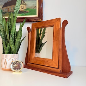 May include: A wooden vanity mirror with a rectangular frame and a warm, reddish-brown finish. The mirror is supported by two curved side pieces and sits on a base. A small, round clock and a potted plant are nearby.
