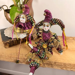 May include: Two decorative jester figurines with porcelain faces, wearing purple and gold costumes. The jesters have tall, pointed hats and are adorned with sequins and gold trim. One holds a staff with a decorative tassel.