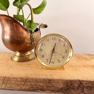 May include: A vintage gold-coloured Big Ben alarm clock with a cream-coloured face and black numerals. The clock sits on a wooden surface next to a copper-coloured planter with green leafy plants.