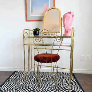 May include: A vintage vanity set with a gold-toned metal frame, featuring a mirror, a small stool with a rust-colored seat, and a decorative pink bust. The set is placed on a black and white geometric patterned rug.