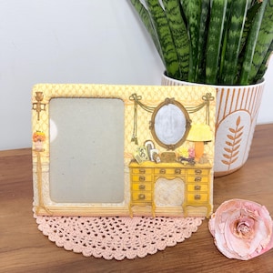 May include: A decorative photo frame in yellow and cream. The frame depicts a miniature vanity scene with a mirror, lamp, and drawers. It sits on a pink lace doily, with a potted plant and a decorative flower in the background.