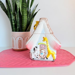May include: A ceramic house-shaped cookie jar with a white roof, pink accents, and a brown speckled trim. The jar features a zebra, hedgehog, and giraffe. The jar sits on a pink mat, with a potted plant in the background.