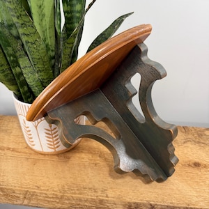 May include: A wooden wall shelf with a dark green and brown finish. The shelf has a curved top and decorative brackets. A potted plant with green leaves is in the background.