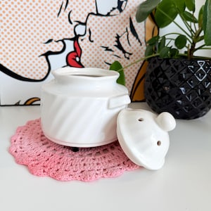 May include: A white ceramic jar with a lid, resting on a pink crocheted doily. The jar has a spiral design and a small handle. The lid has a small knob and two holes. A Pop Art print is in the background.