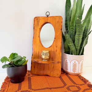 May include: A rustic wooden wall mirror with an oval mirror and a small shelf. A small brass owl figurine sits on the shelf. The mirror is surrounded by a natural wood frame. The background includes plants and an orange woven placemat.