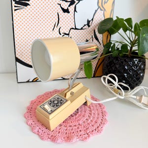 May include: A vintage beige desk lamp with a chrome accent, resting on a pink crocheted doily. The lamp has a rectangular base with a floral design on the switch. A pop art style painting is in the background.