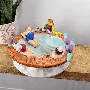 May include: A ceramic serving dish with a spa theme. The dish features a light blue pool with three miniature figures lounging. The rim is brown with a wood-like texture. Two pairs of skis are on the side. The base is white.