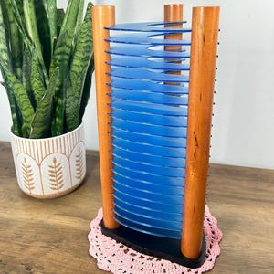 May include: A vintage CD holder with a black base, wooden supports, and blue plastic dividers. The holder is displayed on a pink crocheted doily. A potted snake plant is in the background.