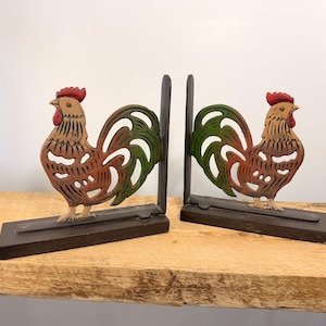May include: A pair of decorative rooster bookends. Each bookend features a rooster with a red comb, brown and orange body, and green tail feathers. The rooster is mounted on a dark brown rectangular base.