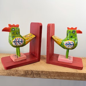 May include: Pair of hand-painted wooden rooster bookends. Each rooster is green with yellow and blue accents, a red comb and beak, and stands on a pink base. The bookends are red and are designed to hold books upright.