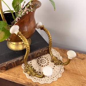 May include: Vintage brass wall hooks with white ceramic knobs and a decorative rope design. The hooks are mounted on a wooden surface, with a small doily and a copper planter with greenery in the background.