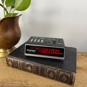 May include: Black and silver Powerhouse Mini Timer alarm clock displaying 12:00 in red digits. The clock sits on a vintage book with gold lettering. A copper planter with greenery is in the background.