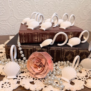 May include: A collection of white seashell-shaped napkin rings with curved handles, arranged on antique books and a lace doily. A pink rose and a strand of faux pearls add decorative elements to the display.