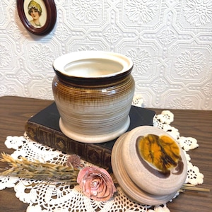 May include: A ceramic jar with a lid, featuring a brown and beige striped design. The lid has a decorative yellow and brown leaf pattern. A small framed portrait hangs on the wall behind the jar.