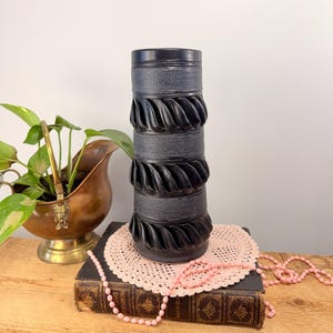May include: A tall, black ceramic vase with a textured, layered design. The vase features alternating bands of smooth and ridged sections. It is displayed on a wooden surface with a vintage book, a pink doily, and a string of pink beads. A brass planter with a green plant is in the background.