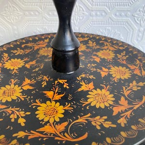 Pu&ograve; includere: Un vassoio decorativo rotondo nero con uno stelo centrale nero. Il vassoio &egrave; adornato con un motivo floreale giallo e arancione. I fiori e le foglie sono dipinti su uno sfondo nero.