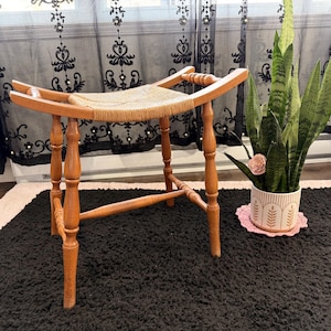 May include: A wooden stool with a woven seat. The stool has four turned legs and a curved top. The seat is made of a light-colored woven material. The stool is set on a black rug.