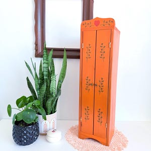 May include: An orange wooden cabinet with two doors, adorned with floral designs and a red heart. The cabinet is placed on a pink doily, with plants and a framed picture in the background. The cabinet is approximately 30 inches tall.