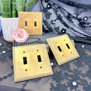 May include: Four vintage brass double light switch covers. Each square cover has two rectangular switch openings and a decorative rope-like border. The brass has a warm, aged patina. The switches are displayed on a dark surface.