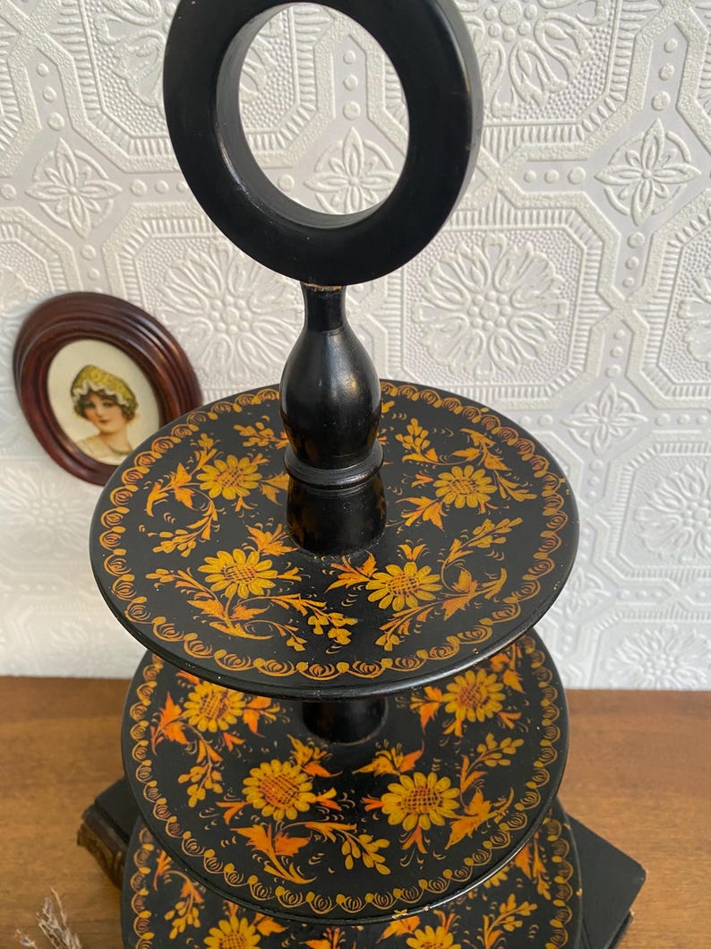 Pu&ograve; includere: Un vassoio nero a tre livelli con manico. Ogni livello &egrave; decorato con un motivo floreale giallo. Il vassoio &egrave; in legno ed &egrave; probabilmente vintage o antico.