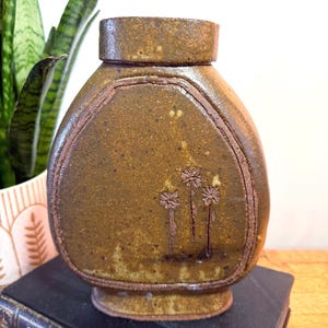 May include: A brown ceramic vase with a textured surface and a decorative floral design. The vase has a rounded body, a flat top, and a base. The vase is approximately 7 inches tall.