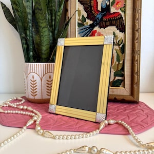 May include: A gold-coloured picture frame with silver detailing, on a pink mat. A pearl necklace and a framed needlepoint artwork are also visible. A potted plant is in the background.