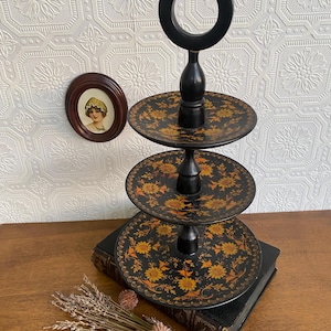 Pu&ograve; includere: Un vassoio nero a tre livelli con manico. Ogni livello presenta un motivo floreale giallo e arancione. Il vassoio &egrave; esposto su una superficie di legno con un libro d'epoca e fiori secchi.