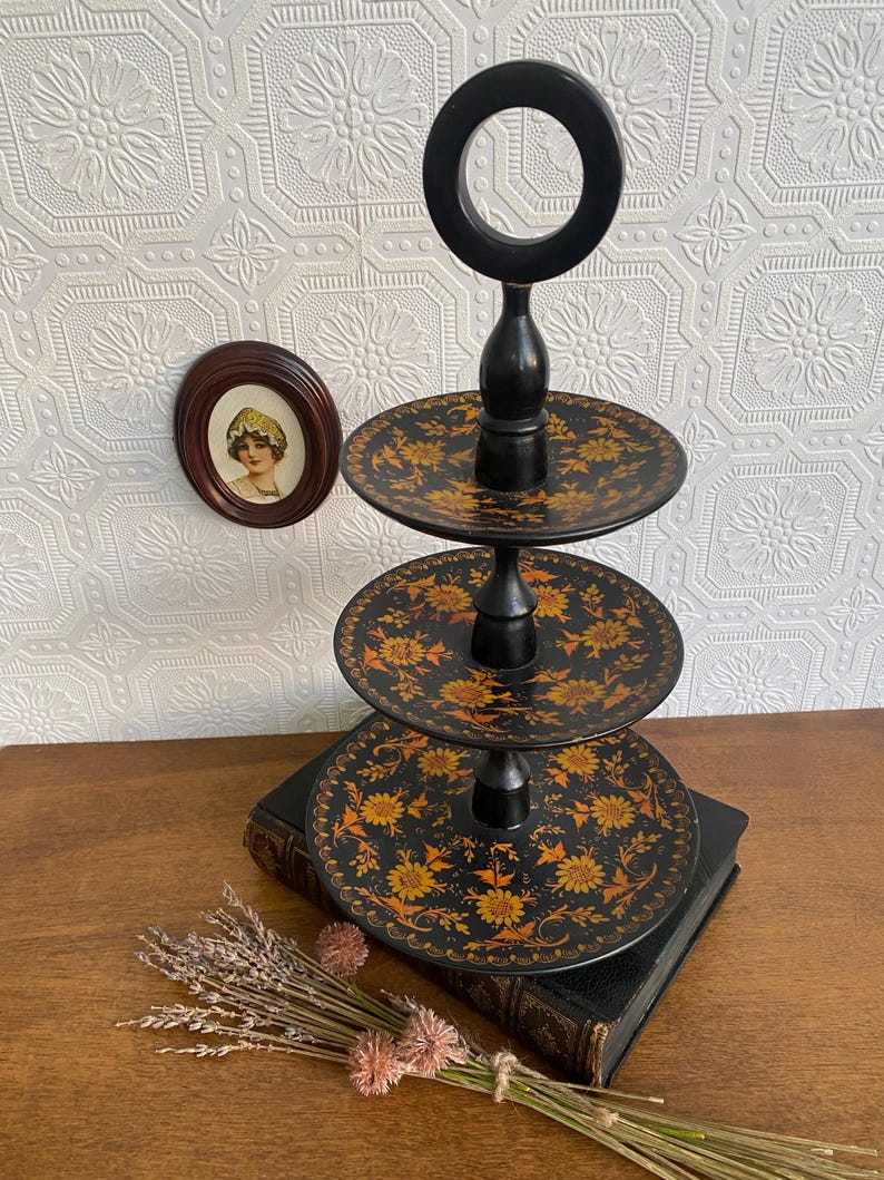 Pu&ograve; includere: Un vassoio nero a tre livelli con manico. Ogni livello presenta un motivo floreale giallo e arancione. Il vassoio &egrave; esposto su una superficie di legno con un libro d'epoca e fiori secchi.