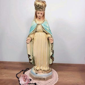 May include: A religious statue of a woman wearing a gold crown and a light blue cape over a cream-coloured dress. The statue is on a pedestal with the text "NOTRE-DAME DU CAP". A pink doily and a black beaded necklace are in front of the statue.