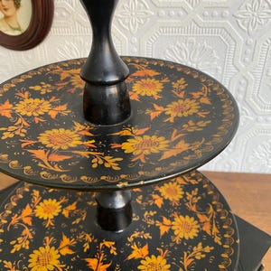 Pu&ograve; includere: Un vassoio a tre livelli con base nera e design floreale. I vassoi sono rotondi e decorati con fiori gialli e arancioni su sfondo nero. Il supporto &egrave; nero e ha un design classico.
