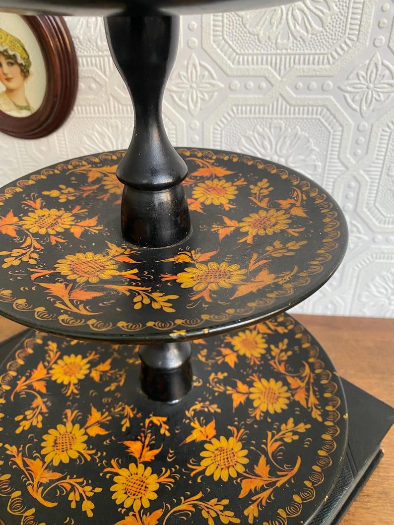 Pu&ograve; includere: Un vassoio a tre livelli con base nera e design floreale. I vassoi sono rotondi e decorati con fiori gialli e arancioni su sfondo nero. Il supporto &egrave; nero e ha un design classico.