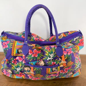 May include: A large, purple tote bag with a vibrant tropical print featuring pink flowers, green leaves, and cartoon characters. The bag has a purple zipper, handles, and trim. The bag is sitting on a wooden surface.