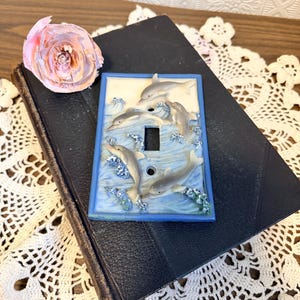May include: A decorative light switch cover featuring a relief of dolphins swimming in blue waves. The cover has a blue border and a neutral background. A pink paper flower and a vintage book are also in the image.