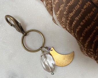 May include: A gold crescent moon charm with a clear crystal bead and purple beads hangs from a brass key ring.