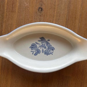 Puede incluir: Plato ovalado para servir con base color crema y diseño floral azul en el centro. El borde está delineado con un fino borde azul. El plato tiene asas en cada extremo.