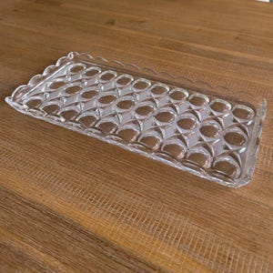 May include: A clear, rectangular glass serving tray with a scalloped edge. The tray features a repeating pattern of oval shapes. The tray is made of clear glass and is sitting on a wooden surface.