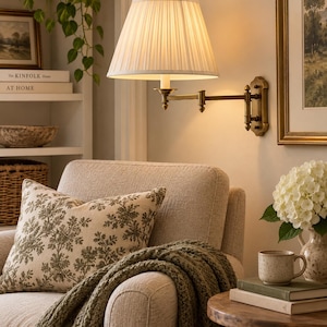 May include: A brass wall-mounted swing arm lamp featuring a pleated off-white fabric shade. The lamp is attached to a rectangular wall plate with an adjustable arm and a small finial at the top of the shade, emitting a warm, soft glow.
