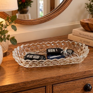 May include: An oval-shaped clear glass tray featuring a textured, geometric diamond-cut pattern along its exterior walls. The tray contains two black electronic car key fobs and a small metal key ring with a black tag attached to the center.