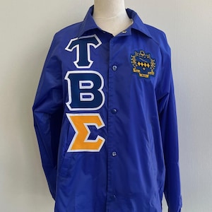 May include: A royal blue jacket with a button-up front and a collared neckline. The jacket features the letters "TBΣ" in white, navy, and gold, along with a crest on the upper right chest. The jacket is displayed on a mannequin.