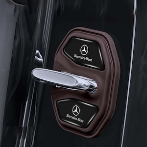 May include: A brown car door handle cover with two black panels featuring the Mercedes-Benz logo and text. The handle is chrome, and the cover is installed on a black car door.