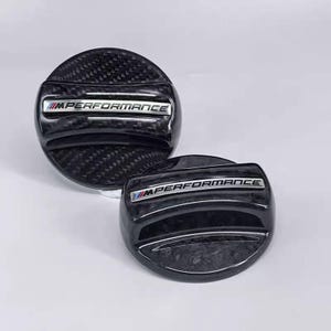 May include: Two black, carbon fibre-textured car accessories. Each has a silver emblem that reads "M PERFORMANCE" in capital letters. The accessories are round with a slightly raised centre and a glossy finish. The background is a solid, light colour.