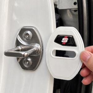 May include: A white car door lock cover with a black and red "S" logo. The cover is being held near the car door lock. The car door lock is silver and has a curved handle.