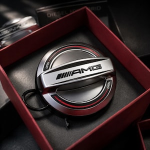May include: A silver and black AMG fuel door cap with red accents, presented in a red box. The cap features the AMG logo and the word "OPEN" on the side. The box is open, revealing the cap.