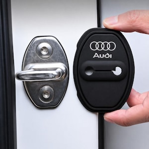May include: Black car door lock cover with the Audi logo in white. The cover is being held next to a silver door latch. The background is a white car door.